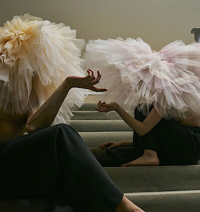 Zwei Tänzer*innen mit Tutus auf dem Kopf sitzen auf einer Treppe mit grauem Teppichbelag